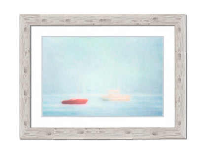 A photo of a small red boat and a small white and yellow boat floating at sea. The photo is edited to have a soft, blurry quality which blends the sky and sea together on the horizon. Printed on paper, matted, and framed.