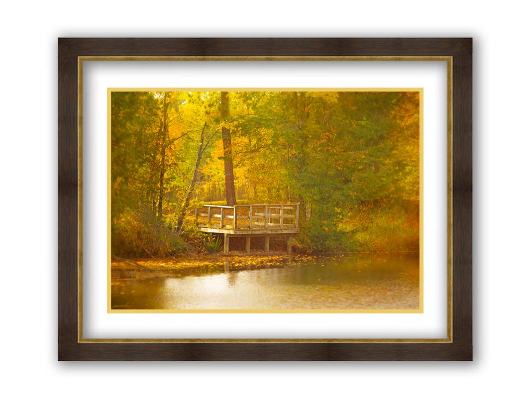 A photo of a small wooden dock, overlooking a forest lake during autumn. Yellow leaves cover the trees and ground, and float atop the water. Printed on paper, matted, and framed.