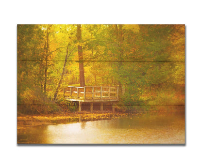 A photo of a small wooden dock, overlooking a forest lake during autumn. Yellow leaves cover the trees and ground, and float atop the water. Printed on a box board.