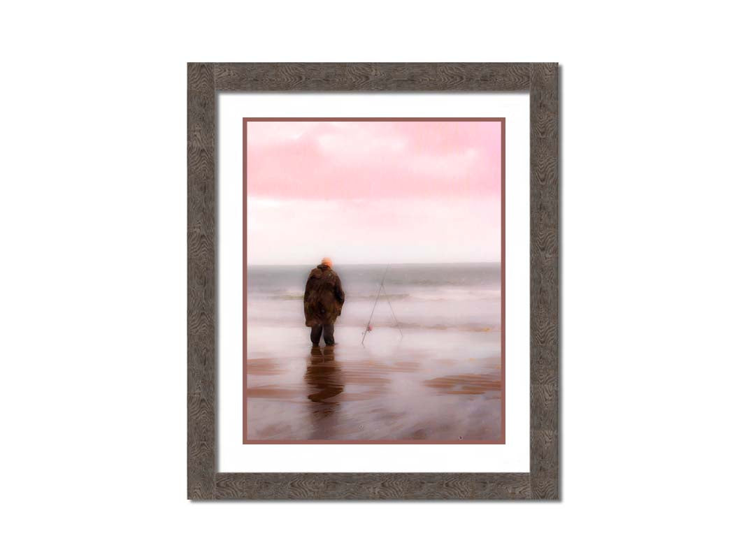 A photograph of a fisherman in oilskins standing in the shallows of the ocean, watching his fishing pole propped up beside him. Printed on paper, matted, and framed.