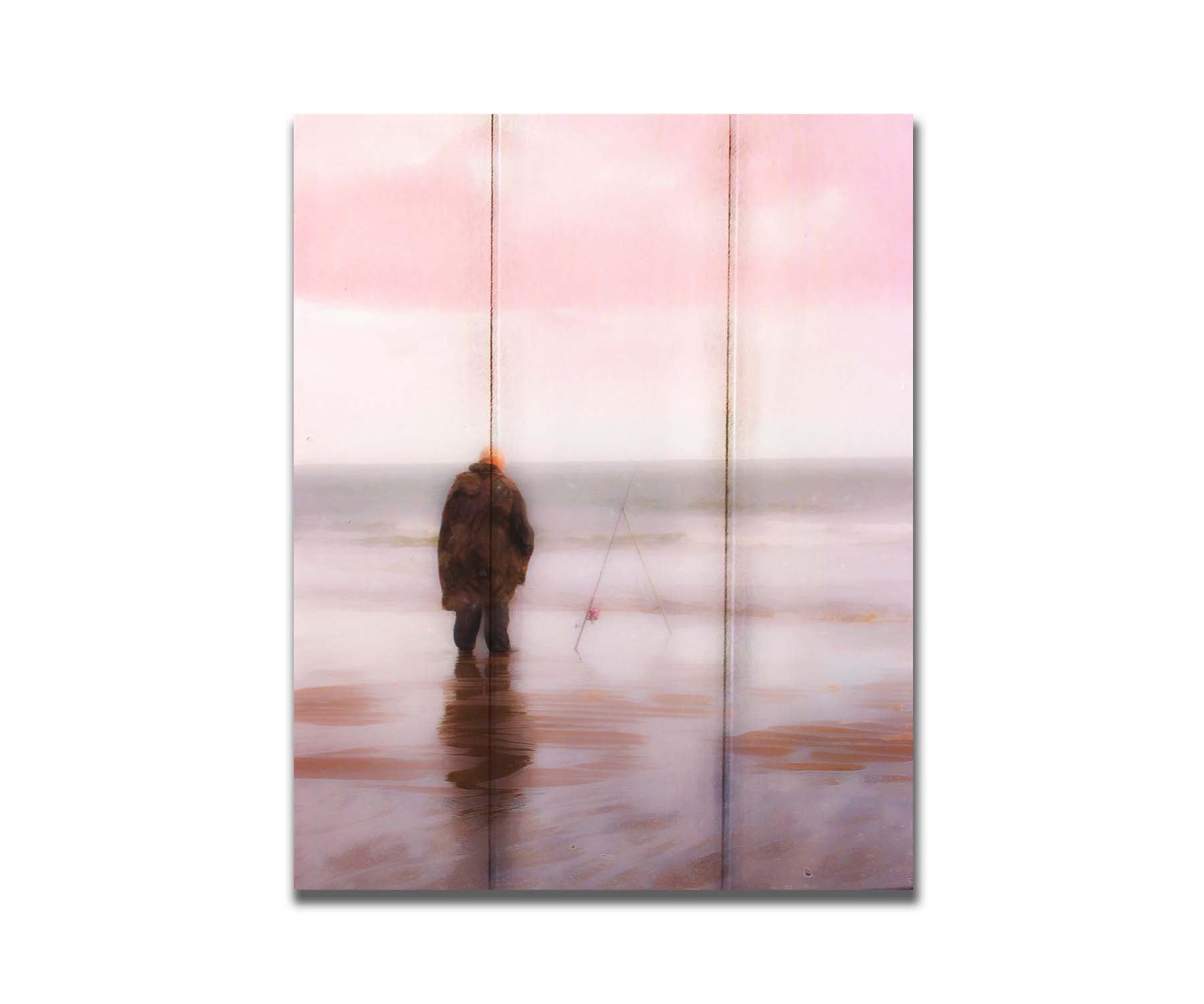 A photograph of a fisherman in oilskins standing in the shallows of the ocean, watching his fishing pole propped up beside him. Printed on a box board.