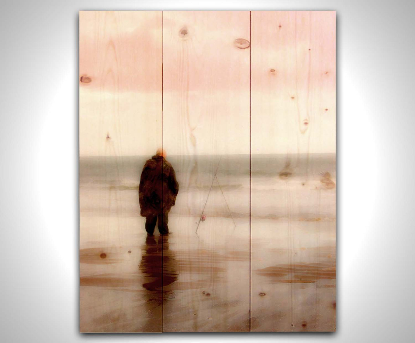 A photograph of a fisherman in oilskins standing in the shallows of the ocean, watching his fishing pole propped up beside him. Printed on a wood pallet.