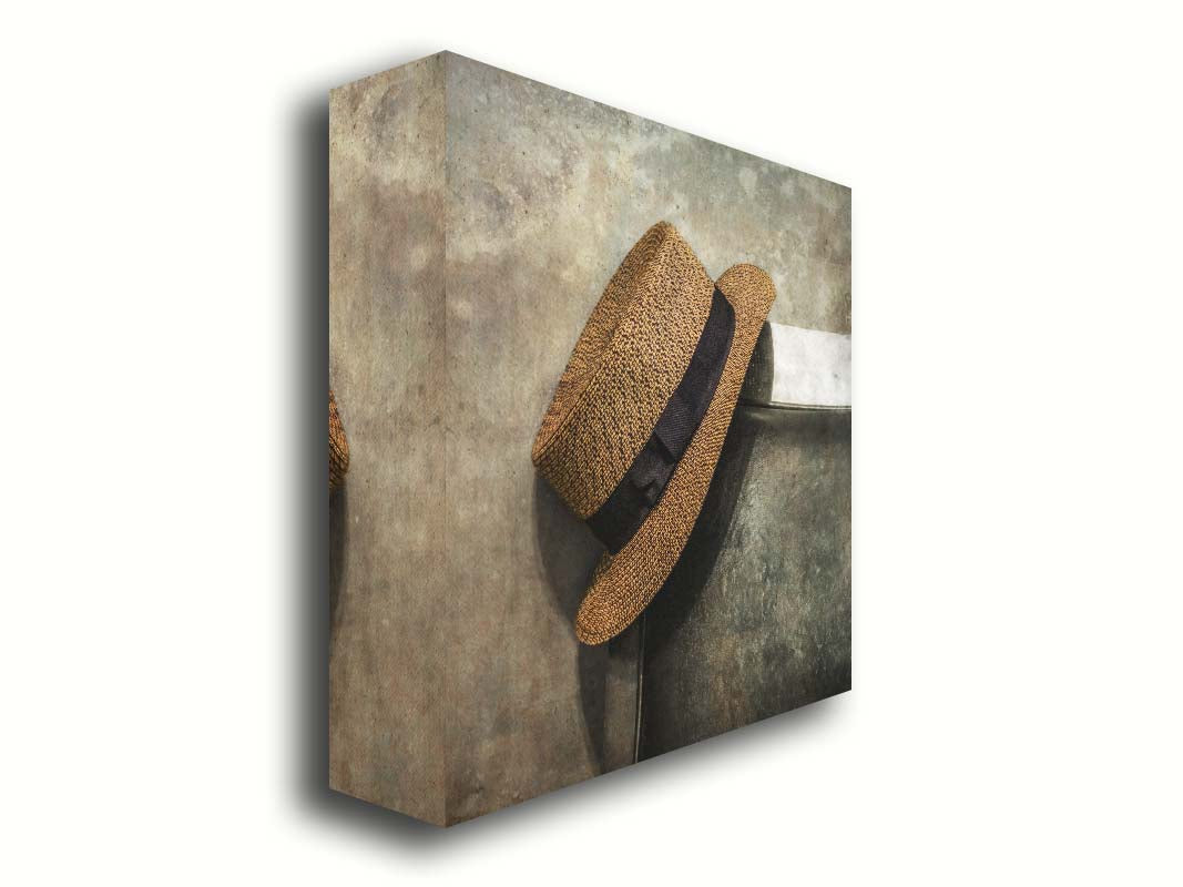 A photograph of a brown boater hat hanging on the back of a chair, against a gray cement wall. Printed on canvas.