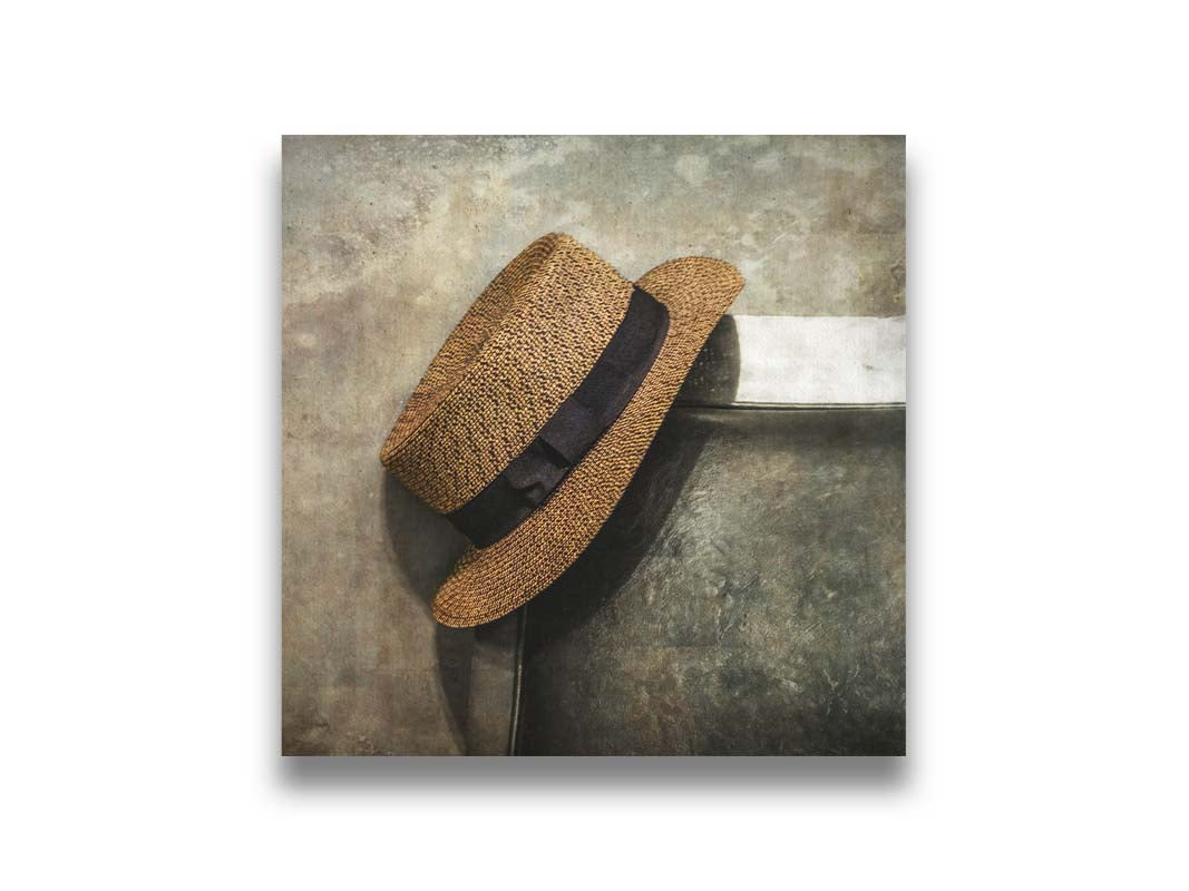 A photograph of a brown boater hat hanging on the back of a chair, against a gray cement wall. Printed on canvas.