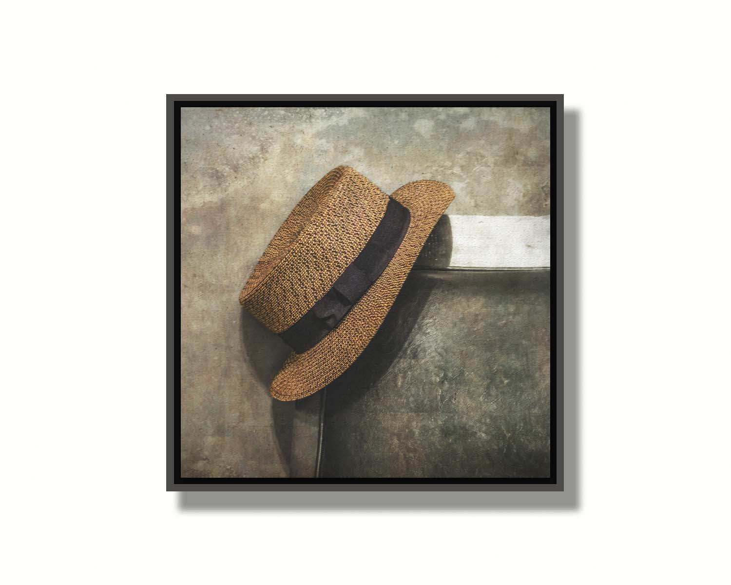 A photograph of a brown boater hat hanging on the back of a chair, against a gray cement wall. Printed on canvas in a float frame.