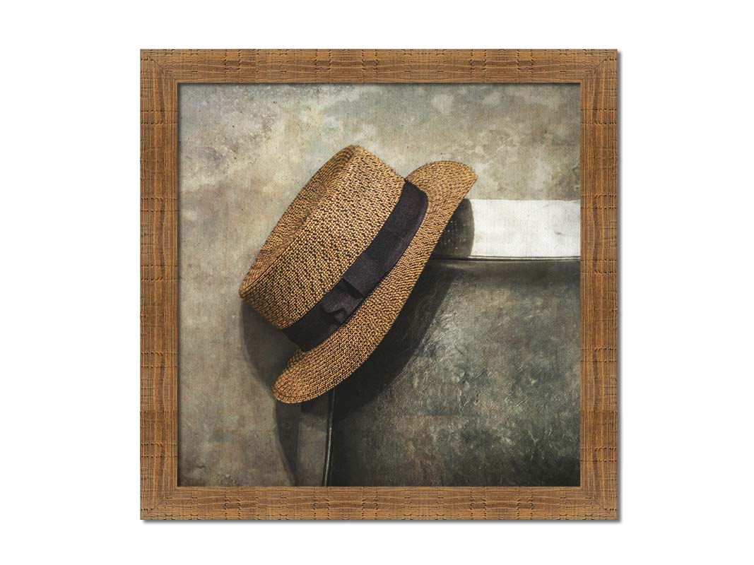 A photograph of a brown boater hat hanging on the back of a chair, against a gray cement wall. Printed on canvas and framed.