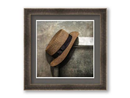 A photograph of a brown boater hat hanging on the back of a chair, against a gray cement wall. Printed on paper, matted, and framed.