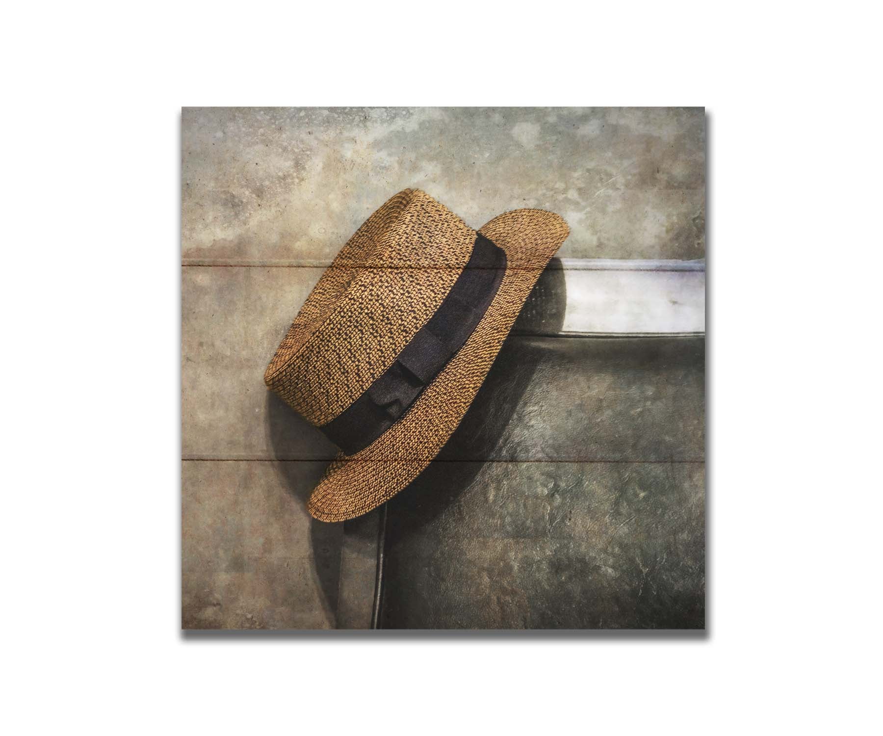 A photograph of a brown boater hat hanging on the back of a chair, against a gray cement wall. Printed on a box board.