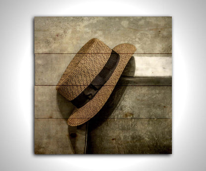 A photograph of a brown boater hat hanging on the back of a chair, against a gray cement wall. Printed on a wood pallet.