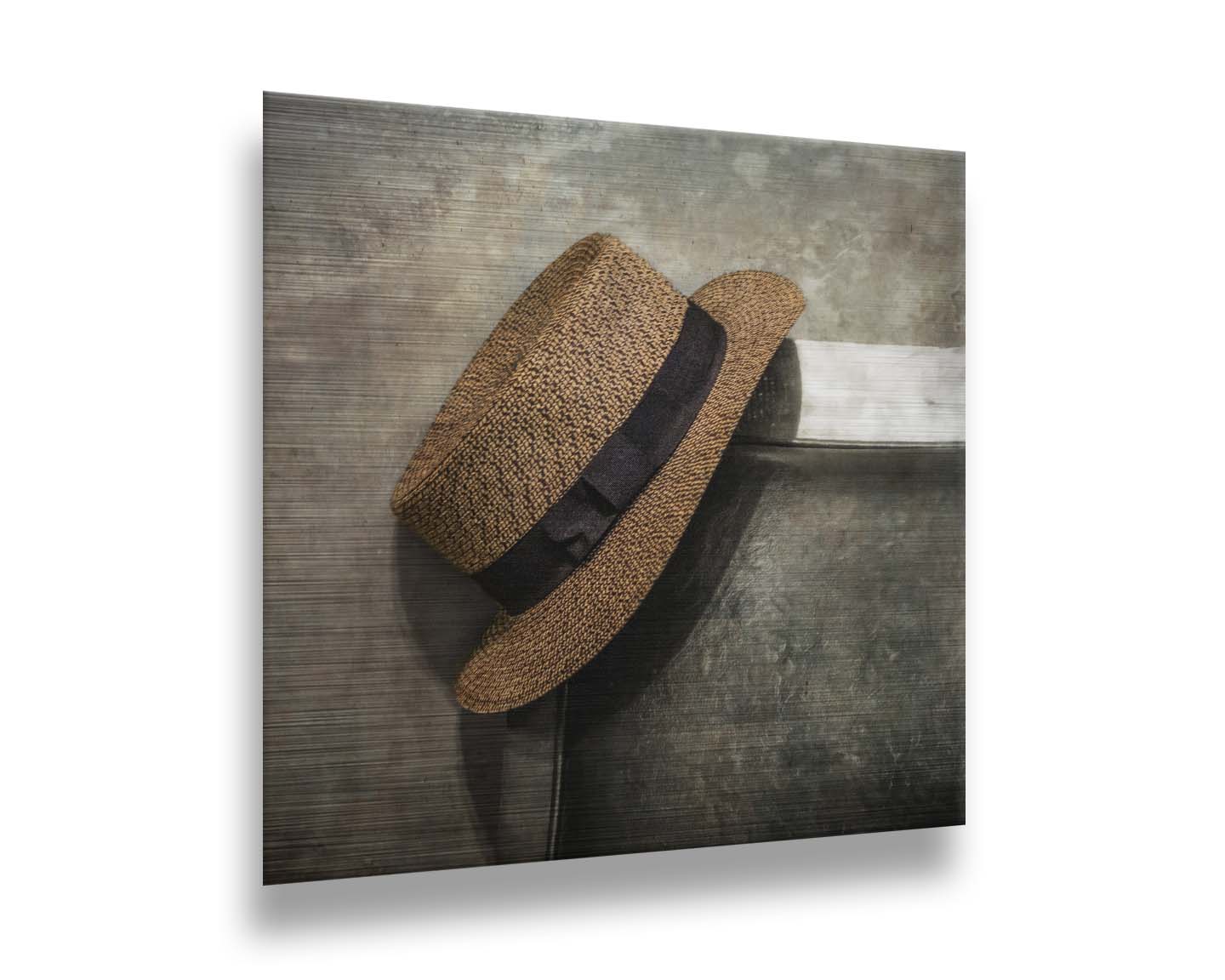 A photograph of a brown boater hat hanging on the back of a chair, against a gray cement wall. Printed on metal.