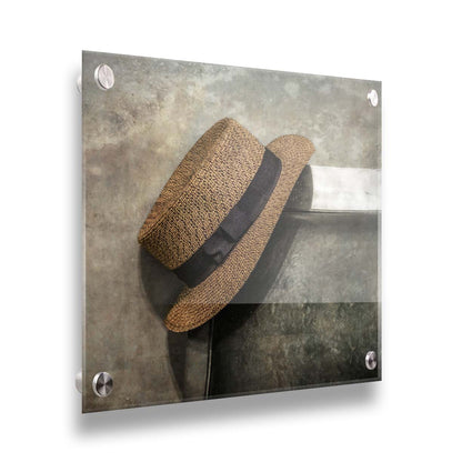 A photograph of a brown boater hat hanging on the back of a chair, against a gray cement wall. Printed on acrylic.