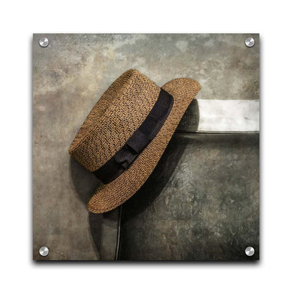 A photograph of a brown boater hat hanging on the back of a chair, against a gray cement wall. Printed on acrylic.