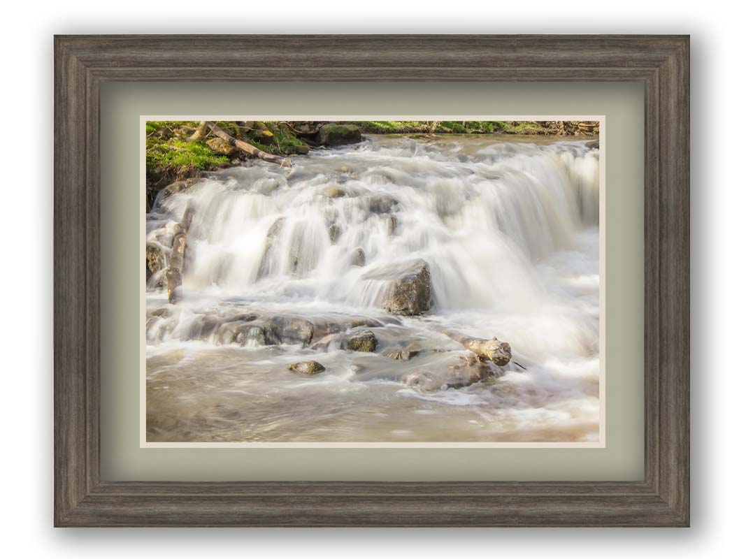 A photograph of a small river waterfall, only a few feet high. The use of a slow shutter speed technique gives the water a soft, gentle appearance. Printed on paper, matted, and framed.