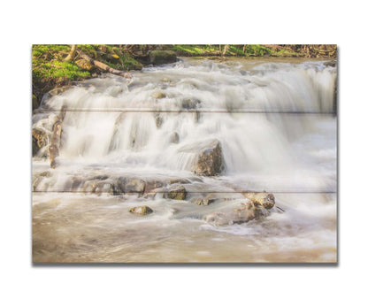 A photograph of a small river waterfall, only a few feet high. The use of a slow shutter speed technique gives the water a soft, gentle appearance. Printed on a box board.