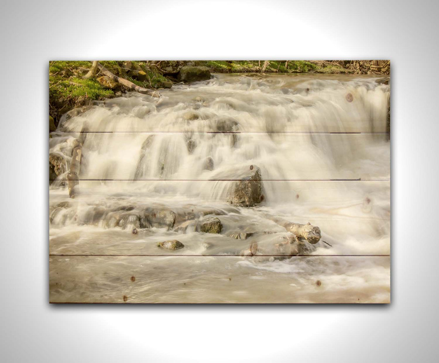 A photograph of a small river waterfall, only a few feet high. The use of a slow shutter speed technique gives the water a soft, gentle appearance. Printed on a wood pallet.