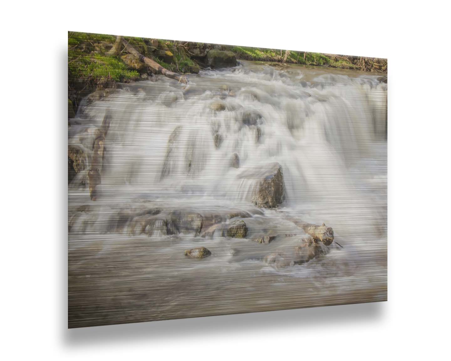 A photograph of a small river waterfall, only a few feet high. The use of a slow shutter speed technique gives the water a soft, gentle appearance. Printed on metal.