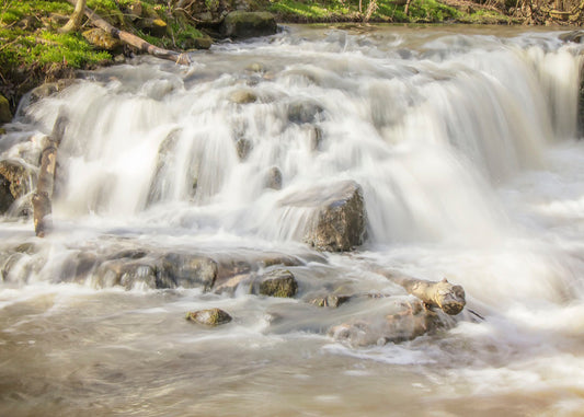 A photograph of a small river waterfall, only a few feet high. The use of a slow shutter speed technique gives the water a soft, gentle appearance. 