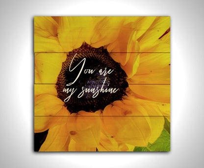 A closeup photo of a bright yellow sunflower, edited in a painterly style and overlaid with the quote "You are my sunshine." Printed on a wood pallet.