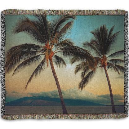 A woven throw blanket designed with a photo of two palm trees in Maui at sunset, with the sea and cloud-topped mountains in the background.