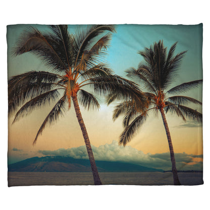 A coral fleece throw blanket designed with a photo of two palm trees in Maui at sunset, with the sea and cloud-topped mountains in the background.
