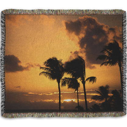 A woven throw blanket designed with a photo of palm trees in Maui silhouetted by the setting sun, with the sea in the background.
