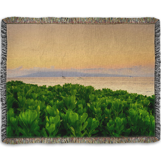 A woven throw blanket designed with a photo overlooking the sea from Kaanapali beach in Hawaii. Green plants fill the foreground, and boats sail on the water in the distance against a gentle orange sky and purple clouds. 