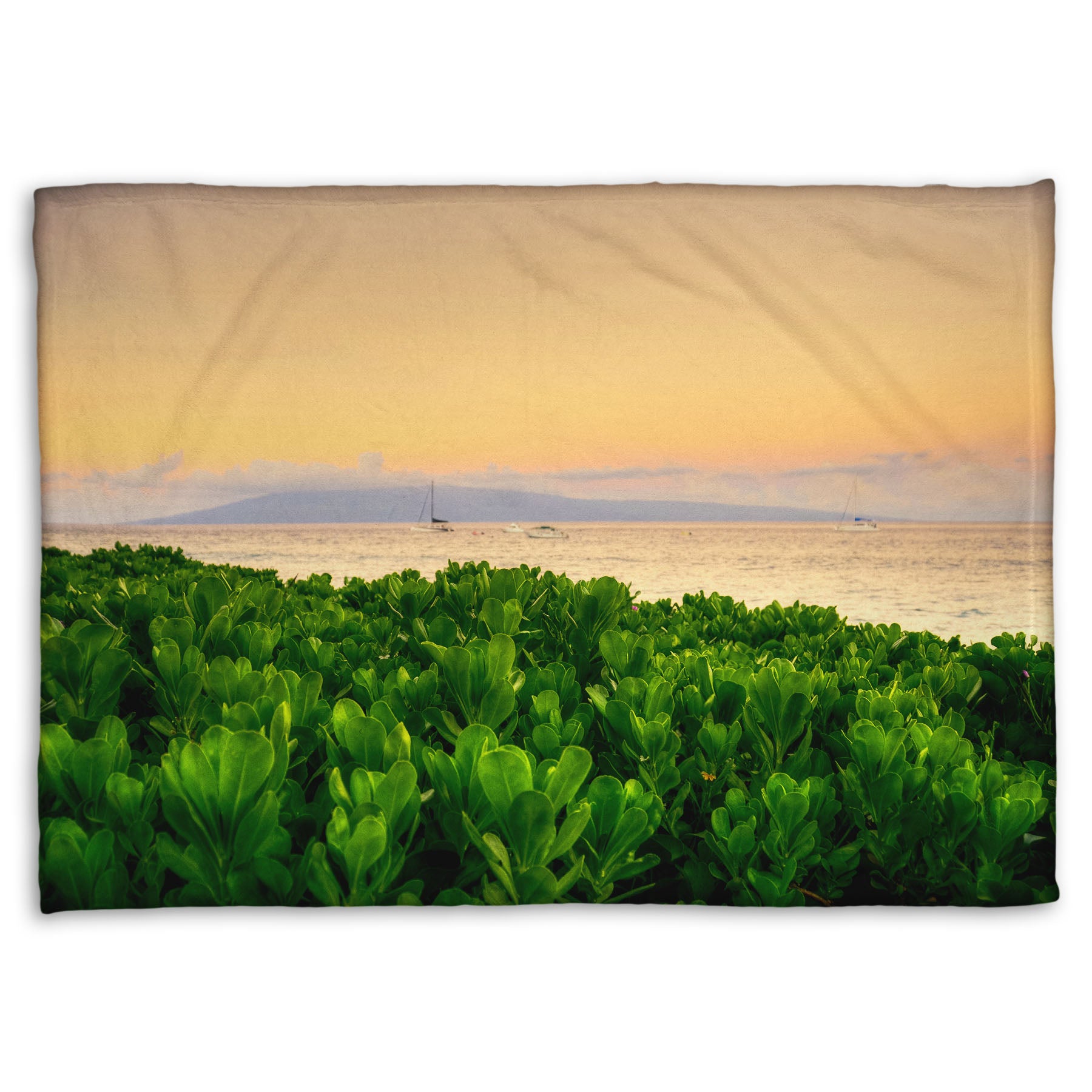 A fleece throw blanket designed with a photo overlooking the sea from Kaanapali beach in Hawaii. Green plants fill the foreground, and boats sail on the water in the distance against a gentle orange sky and purple clouds. 