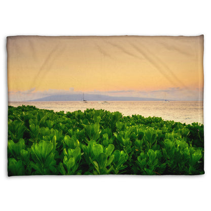 A fleece throw blanket designed with a photo overlooking the sea from Kaanapali beach in Hawaii. Green plants fill the foreground, and boats sail on the water in the distance against a gentle orange sky and purple clouds. 