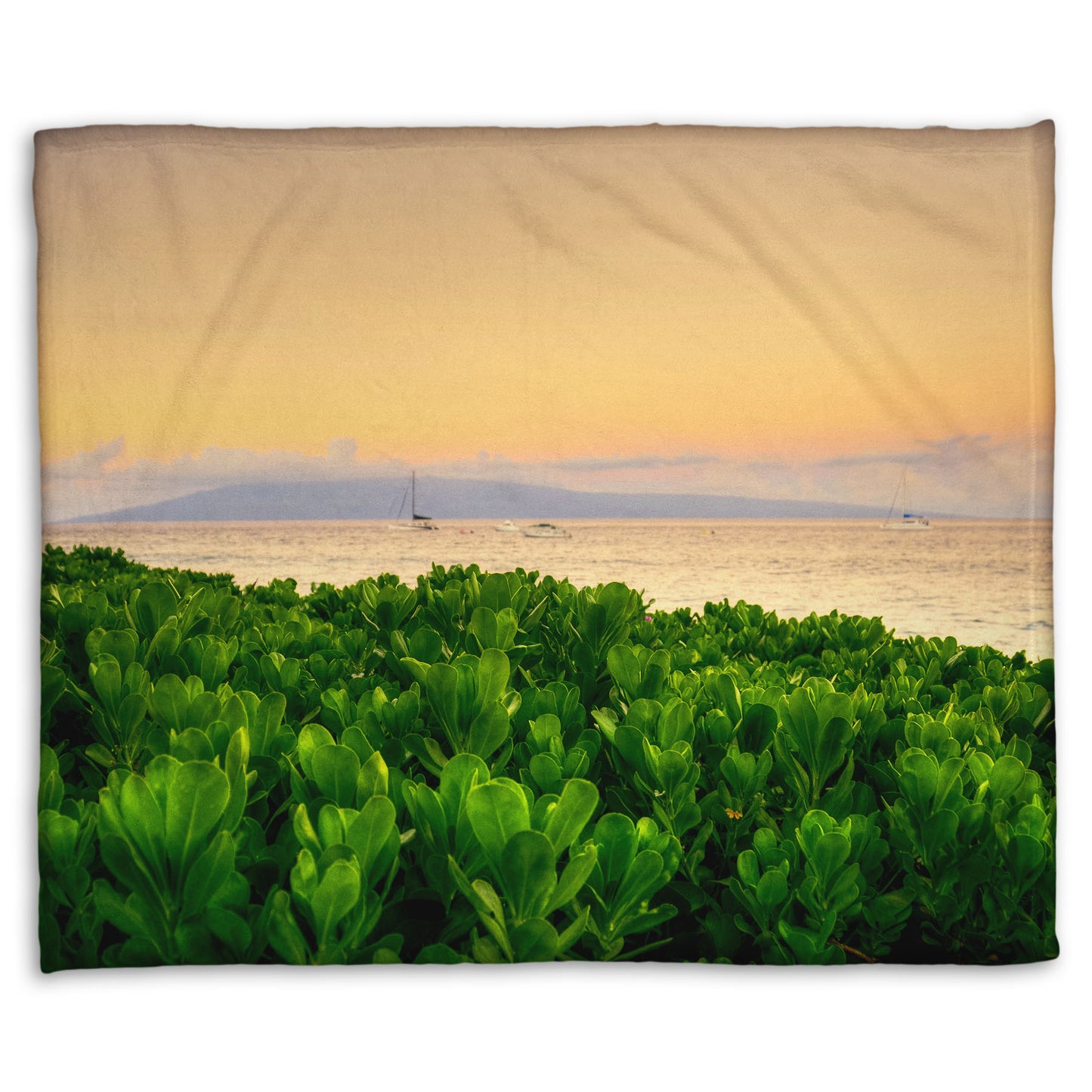 A fleece throw blanket designed with a photo overlooking the sea from Kaanapali beach in Hawaii. Green plants fill the foreground, and boats sail on the water in the distance against a gentle orange sky and purple clouds. 
