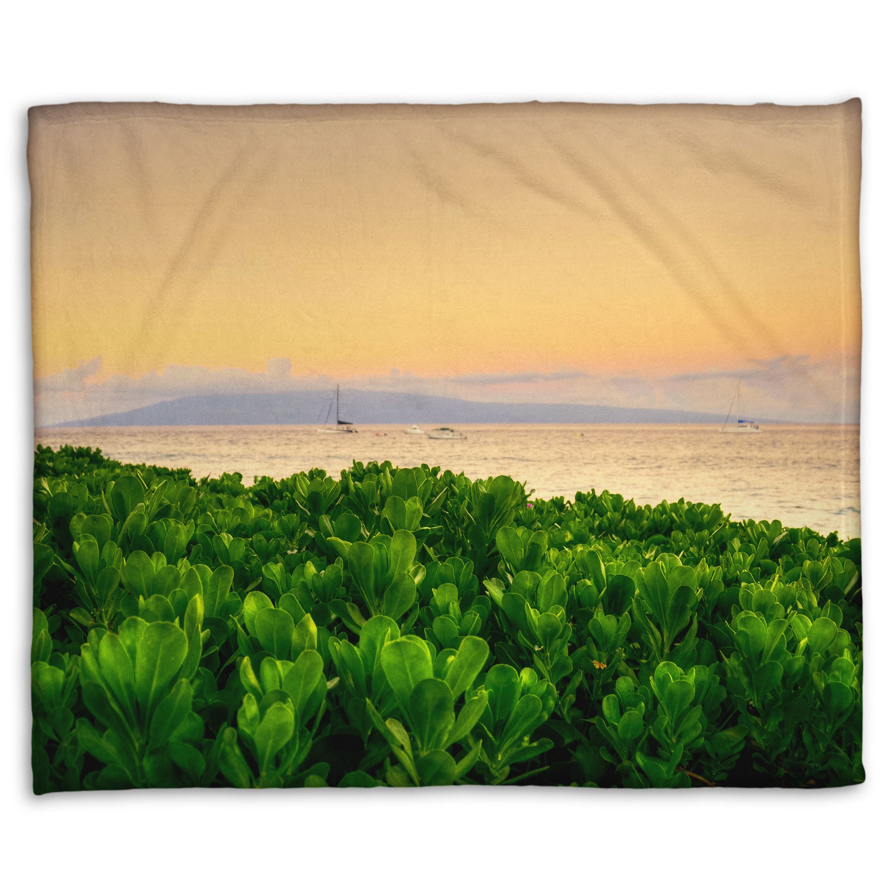 A fleece throw blanket designed with a photo overlooking the sea from Kaanapali beach in Hawaii. Green plants fill the foreground, and boats sail on the water in the distance against a gentle orange sky and purple clouds. 