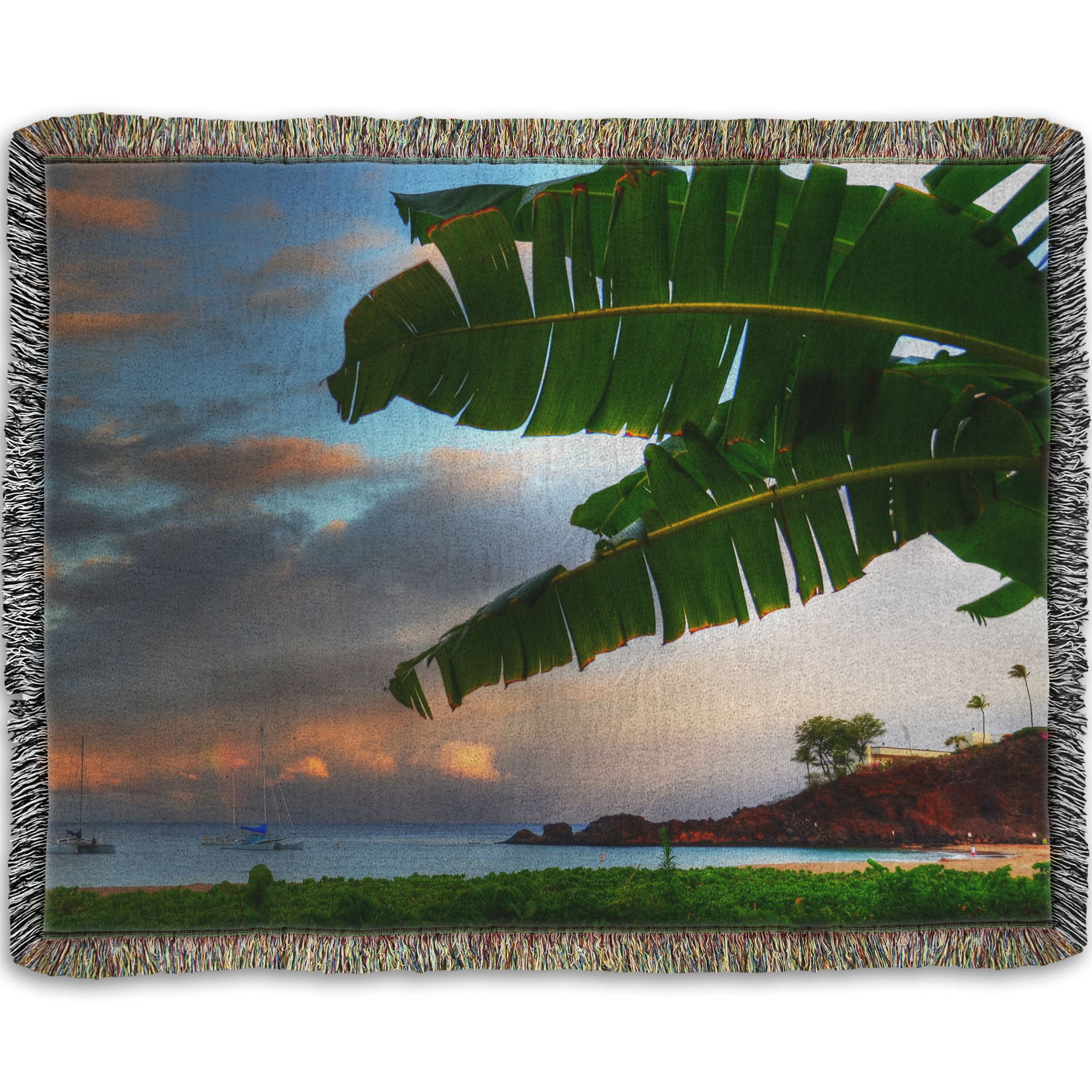 A woven throw blanket designed with a photo of Ka'anapali beach in Hawai'i. Boats can be seen sailing on the ocean, and bright green plants grow in the foreground alongside a palm.