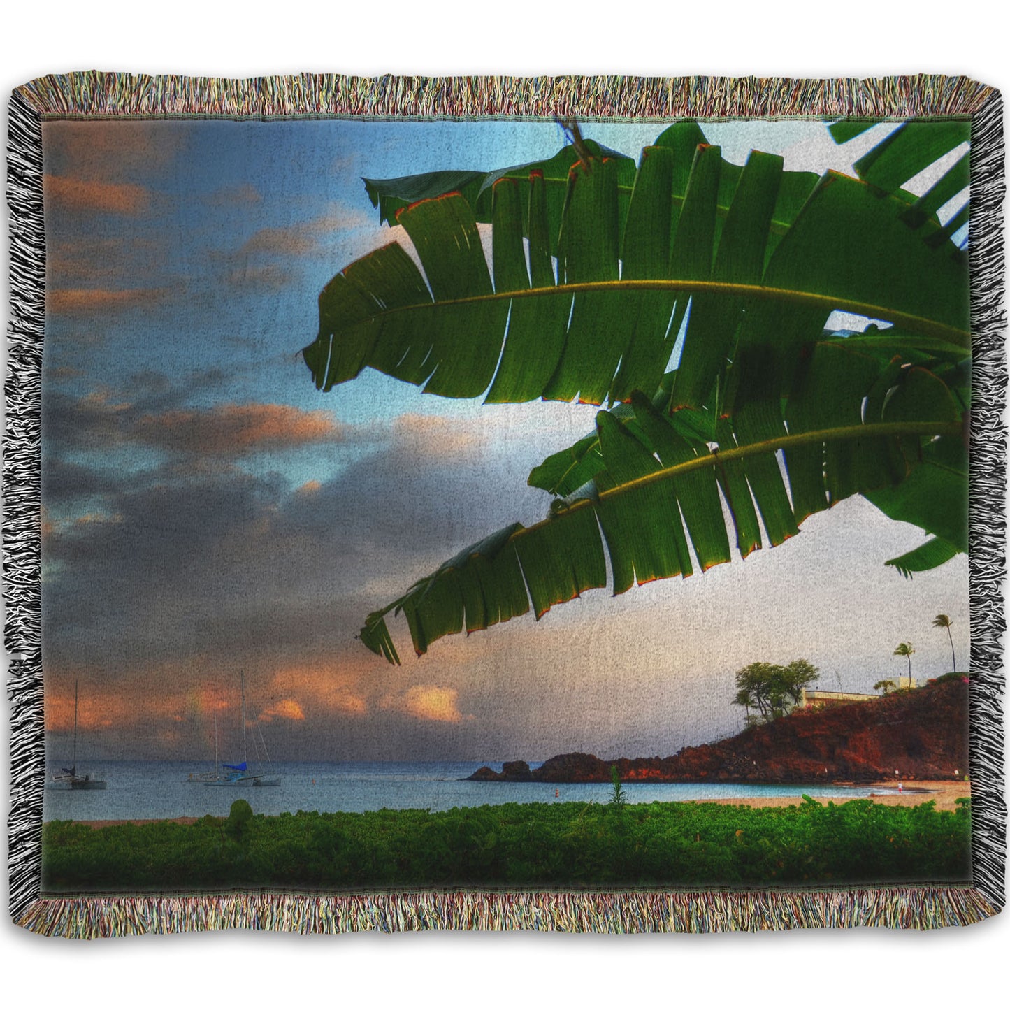 A woven throw blanket designed with a photo of Ka'anapali beach in Hawai'i. Boats can be seen sailing on the ocean, and bright green plants grow in the foreground alongside a palm.