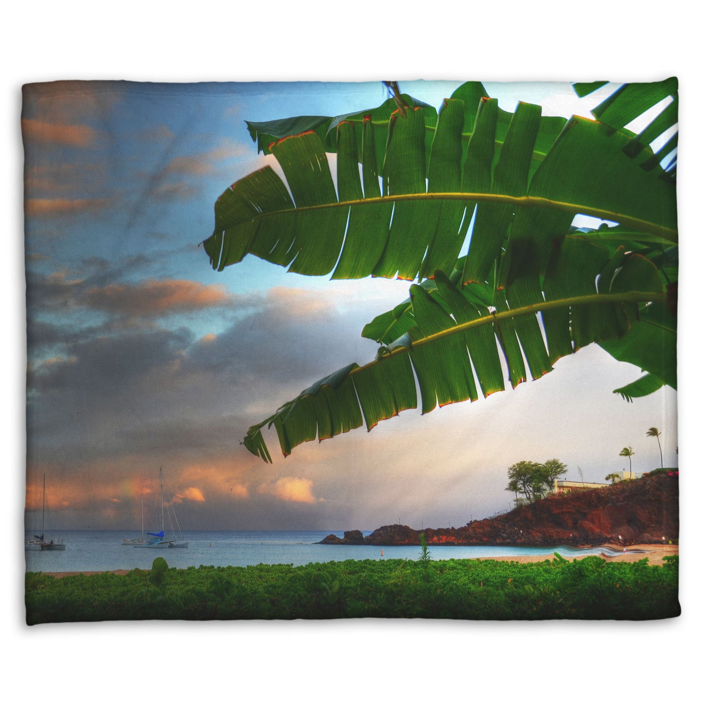 A fleece throw blanket designed with a photo of Ka'anapali beach in Hawai'i. Boats can be seen sailing on the ocean, and bright green plants grow in the foreground alongside a palm.