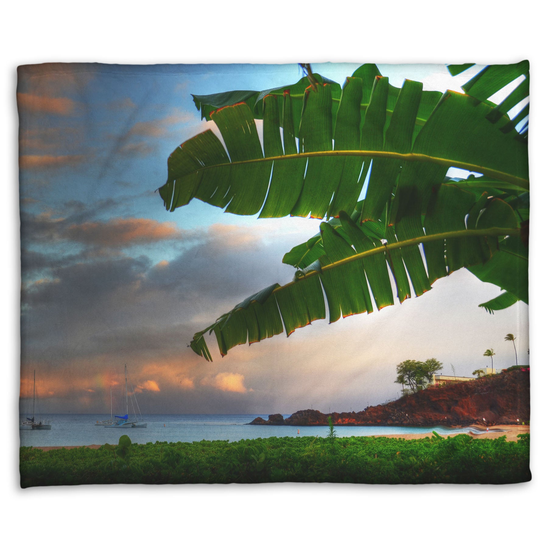A fleece throw blanket designed with a photo of Ka'anapali beach in Hawai'i. Boats can be seen sailing on the ocean, and bright green plants grow in the foreground alongside a palm.