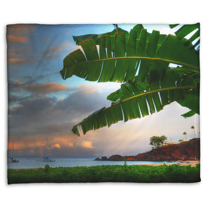 A fleece throw blanket designed with a photo of Ka'anapali beach in Hawai'i. Boats can be seen sailing on the ocean, and bright green plants grow in the foreground alongside a palm.