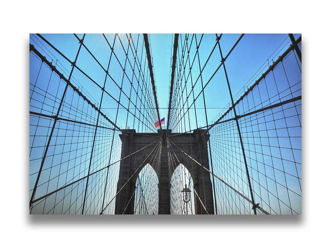 A photo of the Brooklyn Bridge, looking up the suspender cables at one of the towers, topped with an American flag. Printed on canvas.