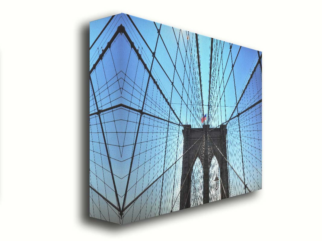 A photo of the Brooklyn Bridge, looking up the suspender cables at one of the towers, topped with an American flag. Printed on canvas.