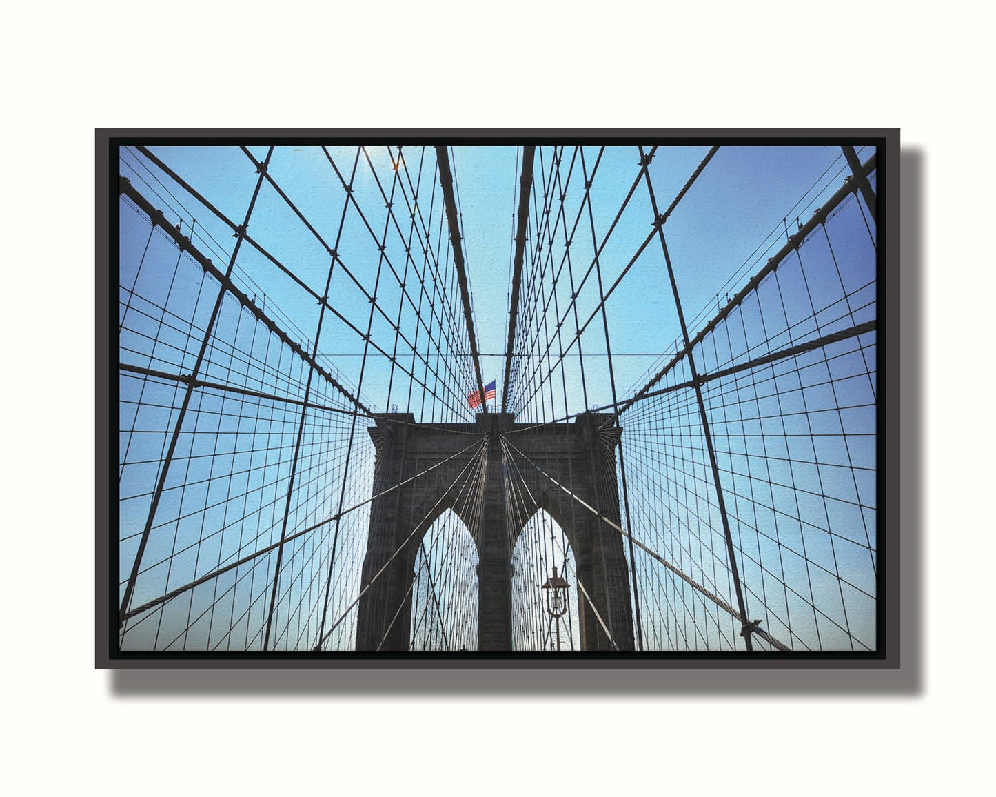 A photo of the Brooklyn Bridge, looking up the suspender cables at one of the towers, topped with an American flag. Printed on canvas in a float frame.