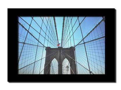 A photo of the Brooklyn Bridge, looking up the suspender cables at one of the towers, topped with an American flag. Printed on canvas and framed.