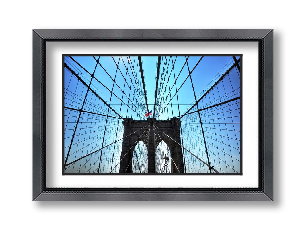 A photo of the Brooklyn Bridge, looking up the suspender cables at one of the towers, topped with an American flag. Printed on paper, matted, and framed.