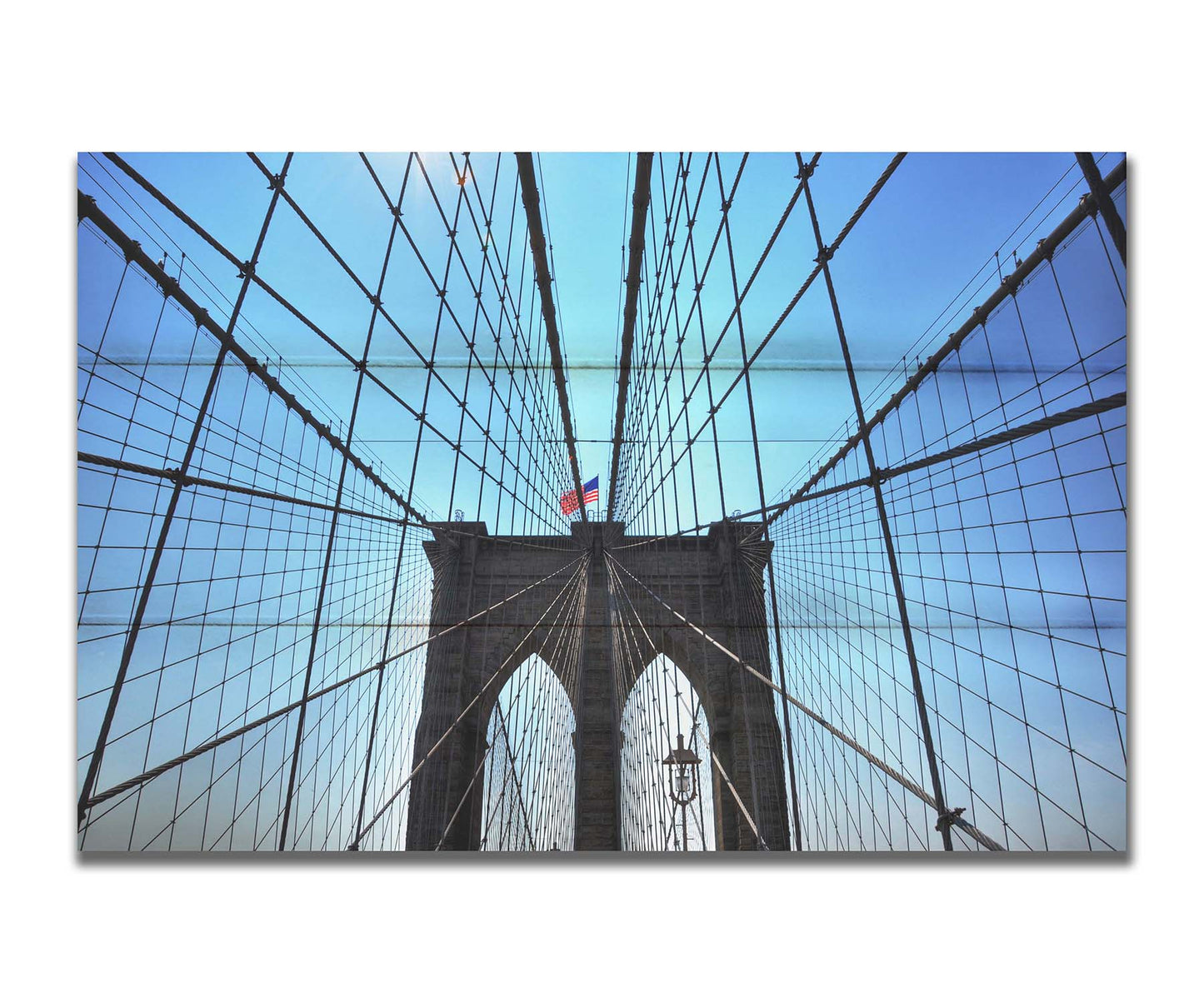 A photo of the Brooklyn Bridge, looking up the suspender cables at one of the towers, topped with an American flag. Printed on a box board.