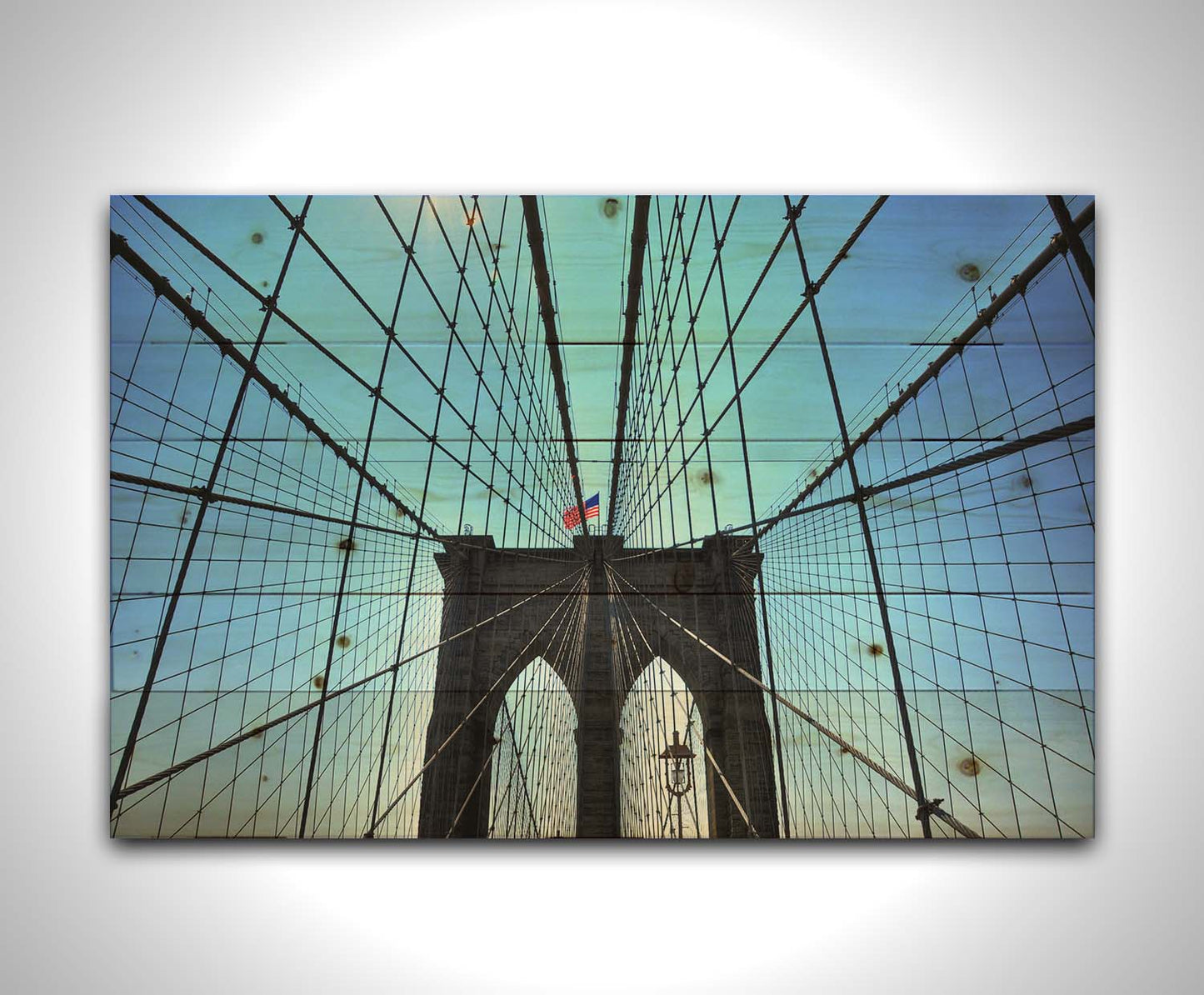 A photo of the Brooklyn Bridge, looking up the suspender cables at one of the towers, topped with an American flag. Printed on a wood pallet.