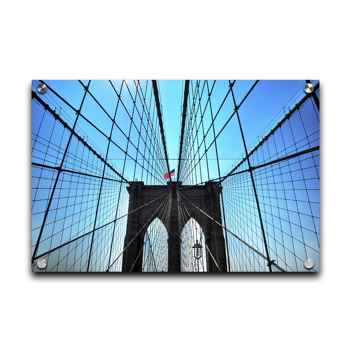 A photo of the Brooklyn Bridge, looking up the suspender cables at one of the towers, topped with an American flag. Printed on acrylic.