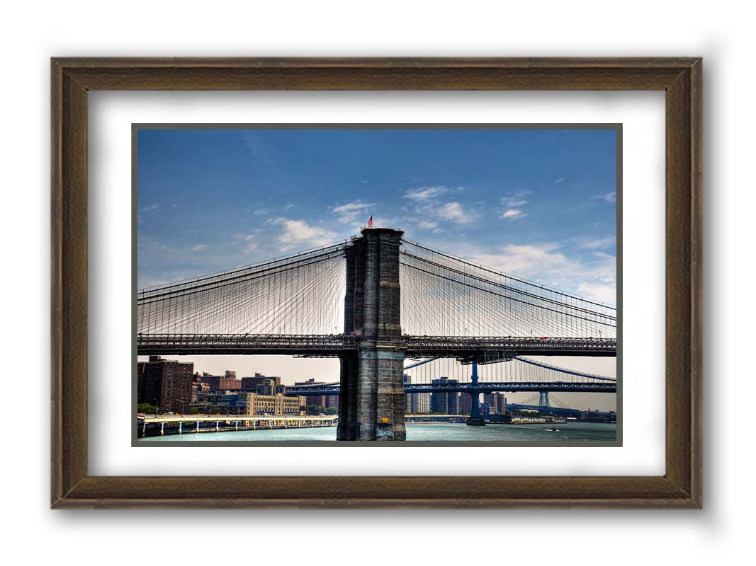 A photo of a side view of the Brooklyn Bridge against a blue sky. Printed on paper, matted, and framed.