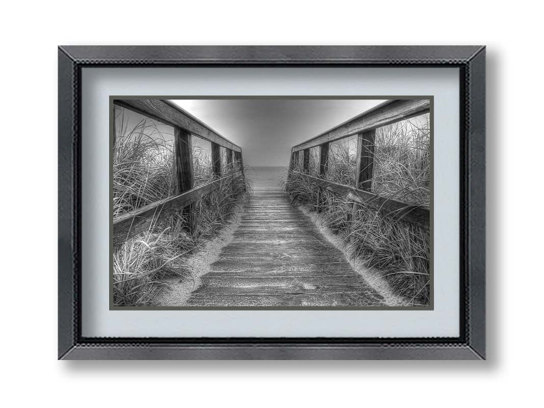 A grayscale photo looking down a wooden railed path on the beach at Cape Cod. Printed on paper, matted, and framed.