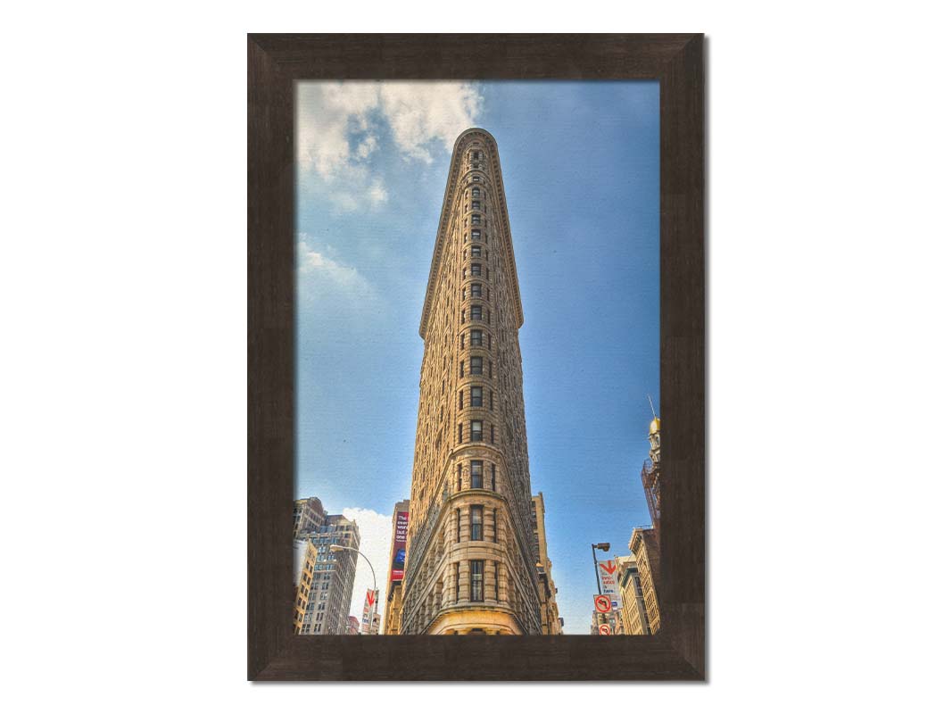 A photo of the Flatiron Building in New York City, New York. Its iconic triangular shape is captures against a bright blue sky. Printed on canvas and framed.