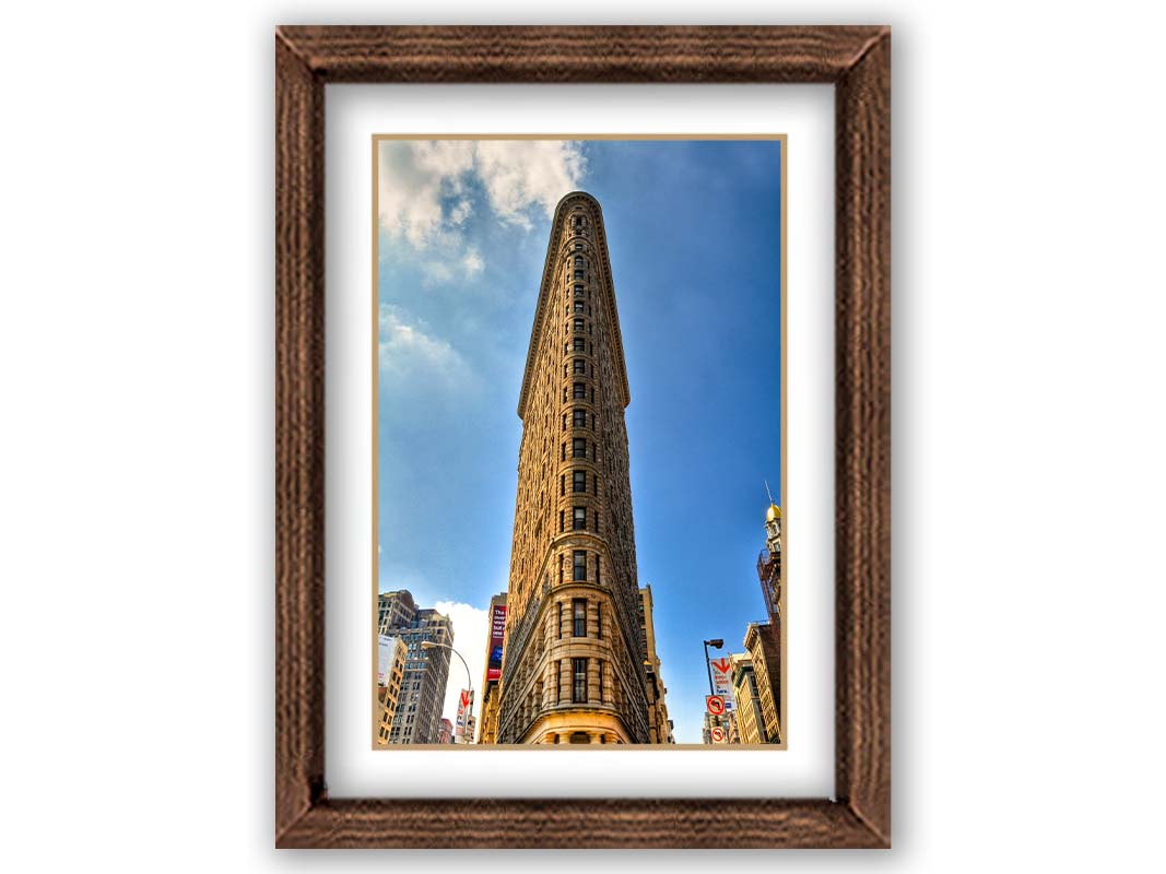 A photo of the Flatiron Building in New York City, New York. Its iconic triangular shape is captures against a bright blue sky. Printed on paper, matted, and framed.