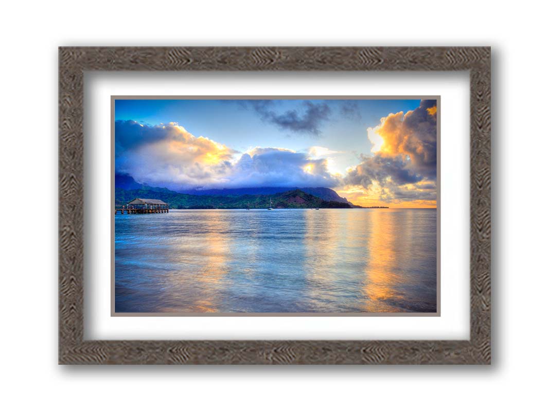 A photo of the ocean at Hanalei Bay at sunset. Clouds rest atop the mountains and reflect the golden light. Printed on paper, matted, and framed.