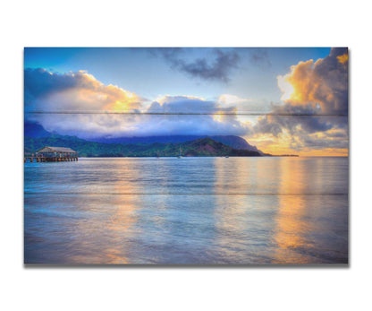 A photo of the ocean at Hanalei Bay at sunset. Clouds rest atop the mountains and reflect the golden light. Printed on a box board.