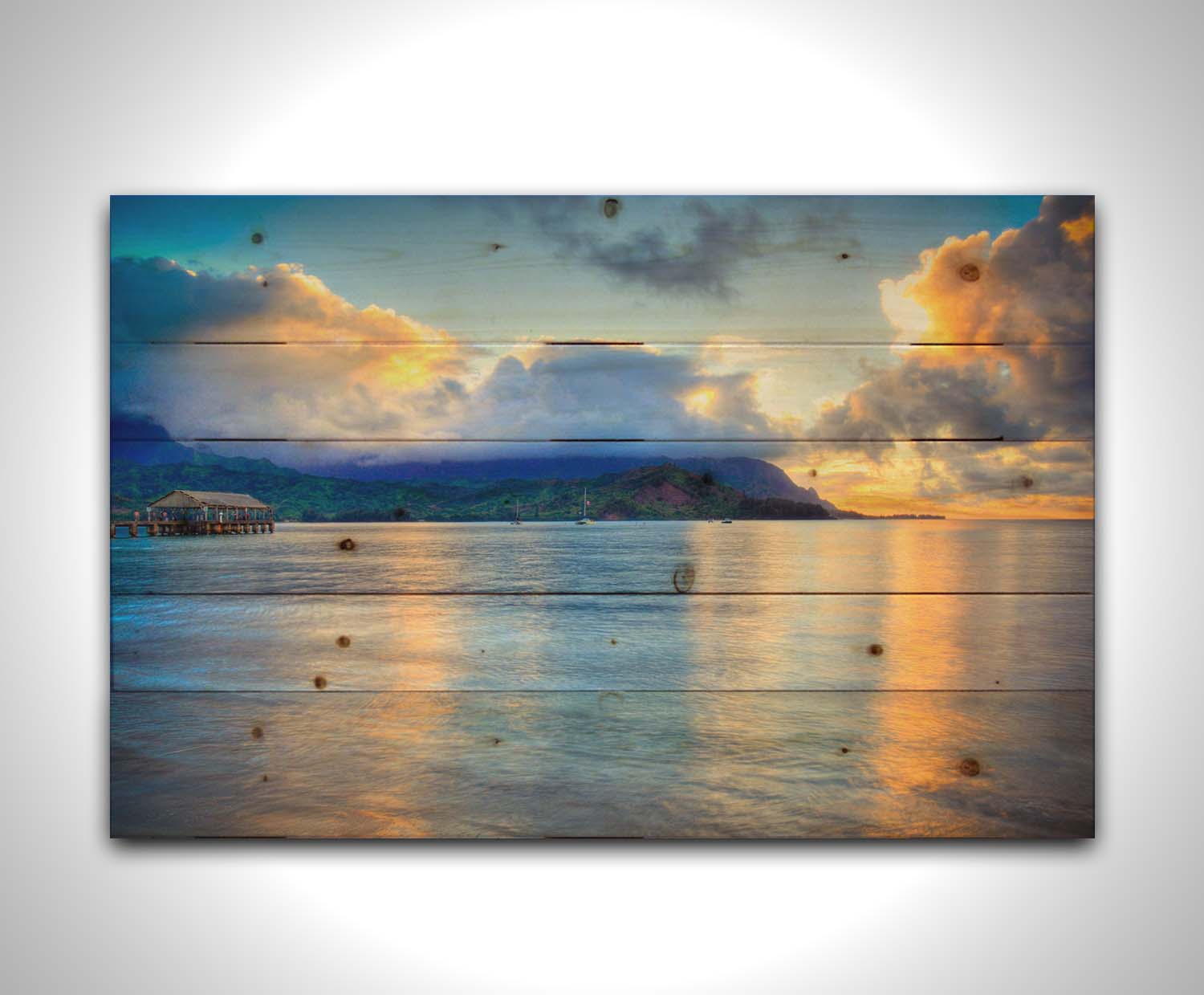 A photo of the ocean at Hanalei Bay at sunset. Clouds rest atop the mountains and reflect the golden light. Printed on a wood pallet.